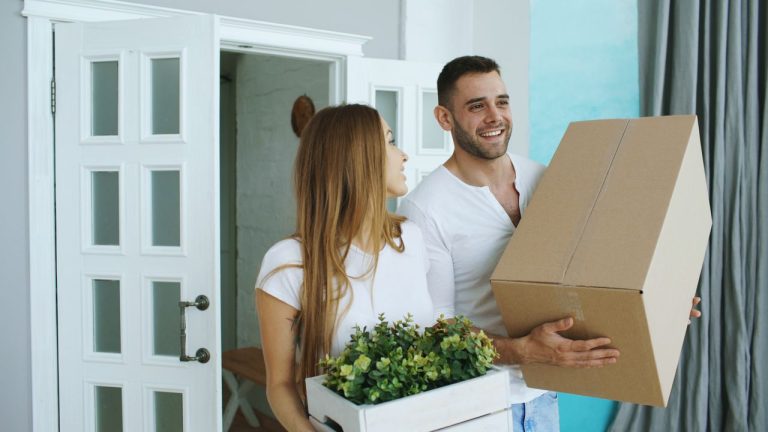 Couple carrying moving boxes and a plant through the front door of a new home